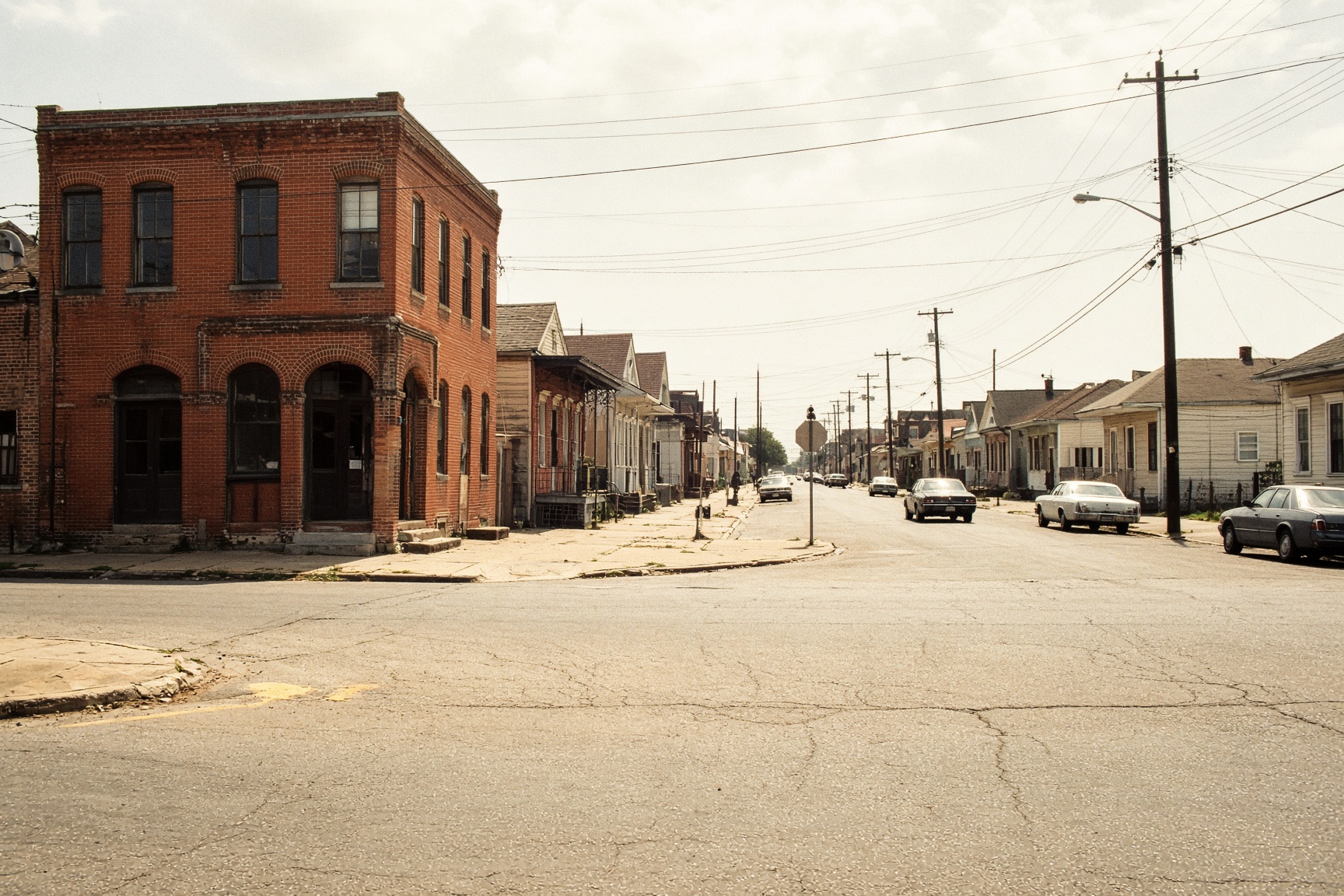 An unshaded New Orleans intersection in harsh midday sun, brick corner store and almost no mature trees.
